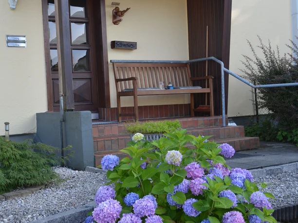 Ferienwohnung Bergisch Land Eingangsbereich eines Hauses mit Holzbänken, Blumen und einer blühenden Hortensie im Vordergrund.