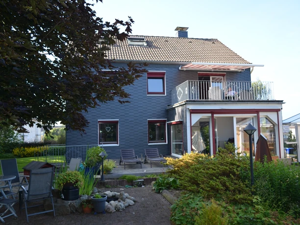 Ferienwohnung Bergisch Land Zweistöckiges Haus mit grauer Fassade, Balkon, Terrassenmöbeln und Garten im Vordergrund.