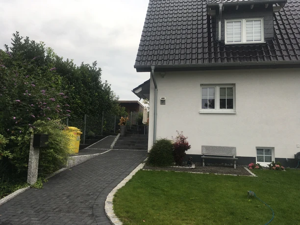 Ferienwohnung Blissenbach Einfamilienhaus mit weißer Fassade, schwarzem Dach, gepflegtem Vorgarten und gepflastertem Weg.