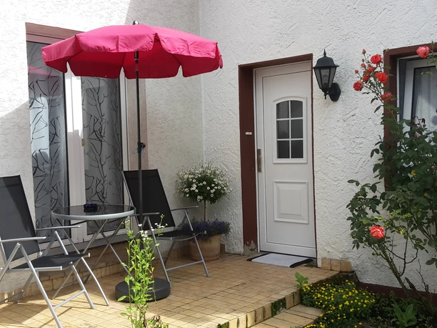 Ferienwohnung Finklenburg Kleiner Hof mit Tisch und Stühlen unter rotem Sonnenschirm vor einer weißen Hausfassade.