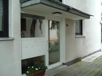 Ferienwohnung Helli Hecht Moderne Eingangstür eines Wohngebäudes mit weißem Rahmen, Glaseinsatz, Briefkästen und Topfpflanze.