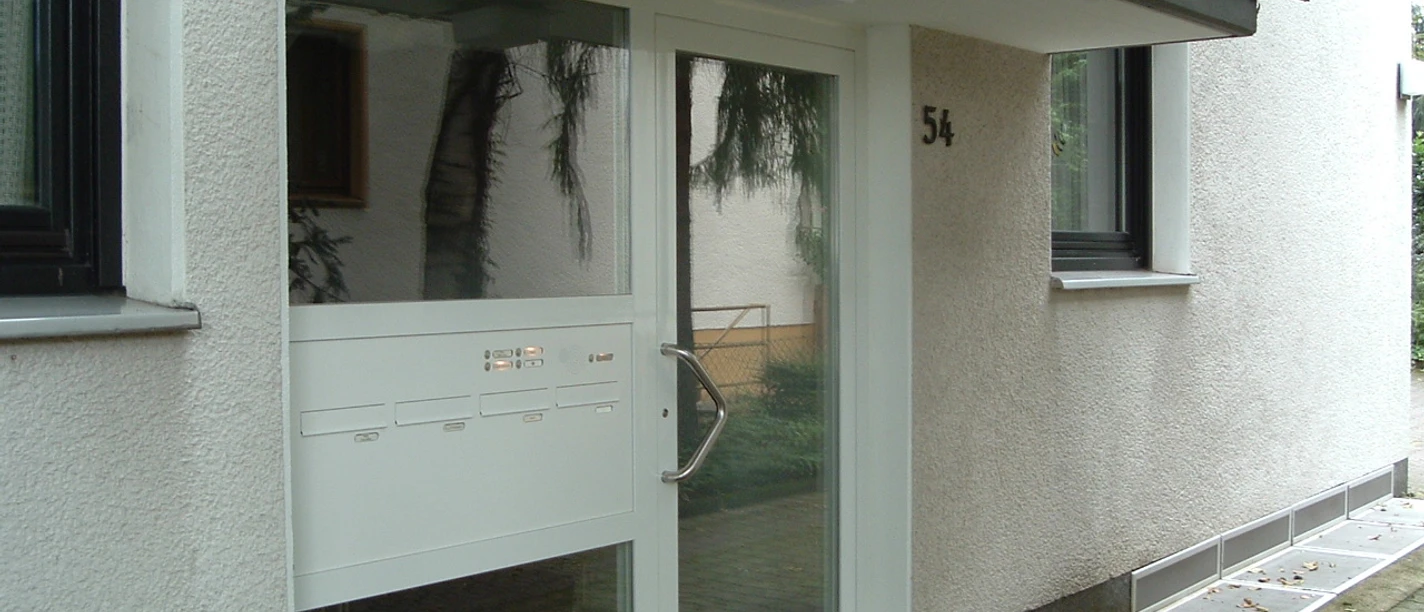 Ferienwohnung Helli Hecht Moderne Eingangstür eines Wohngebäudes mit weißem Rahmen, Glaseinsatz, Briefkästen und Topfpflanze.