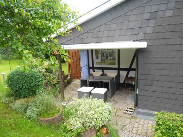 Ferienwohnung Hennemann Sitzbereich im Freien mit Korbmöbeln unter weißem Vordach, umgeben von grüner Vegetation.