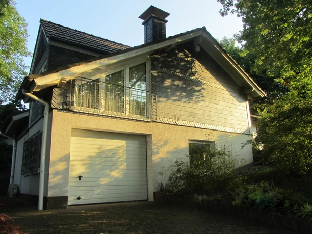 Hauptansicht Aussen Ein Einfamilienhaus mit Dachgaube, großen Fenstern und einer Garage, umgeben von Bäumen im Abendlicht.