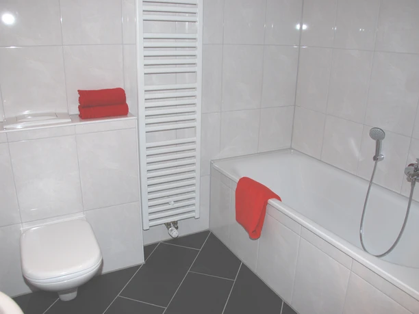 Bad Helles Badezimmer mit Badewanne, WC, Handbrause und roten Handtüchern, weiße Fliesen und Handtuchheizkörper.