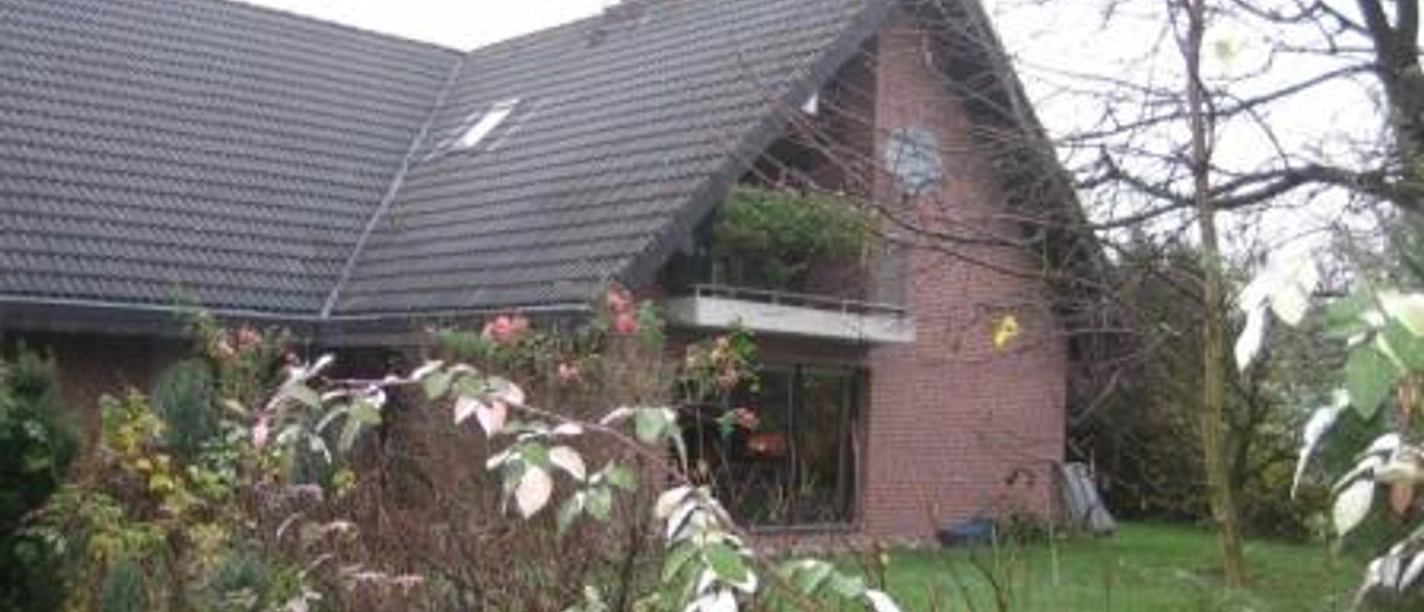 Außenansicht Backsteinhaus mit steilem Satteldach, Balkon und Garten, umgeben von Herbstbäumen und Büschen.