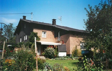 Ferienwohnung Haas Zweistöckiges Backsteinhaus umgeben von üppigen Gärten, blühenden Pflanzen und klarer Sommerhimmel.
