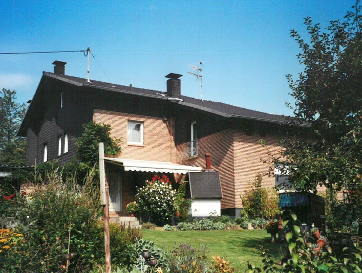 Ferienwohnung Haas Zweistöckiges Backsteinhaus umgeben von üppigen Gärten, blühenden Pflanzen und klarer Sommerhimmel.