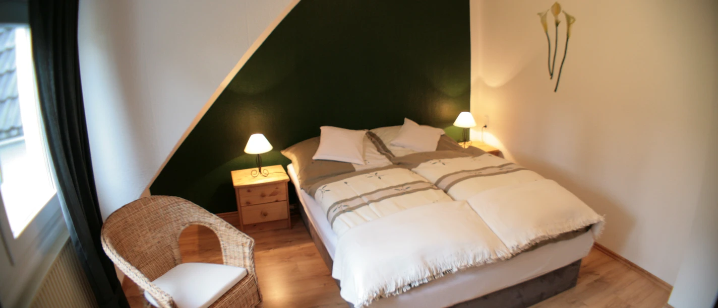 Ferienwohnung Scholz Gemütliches Schlafzimmer mit Doppelbett, zwei Nachttischen, Lampen und einem Korbstuhl.