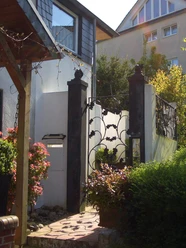 Gartenansicht Zierliches Gartentor aus Metall mit kunstvollem Rankendekor, flankiert von Pflanzen in Sonne.
