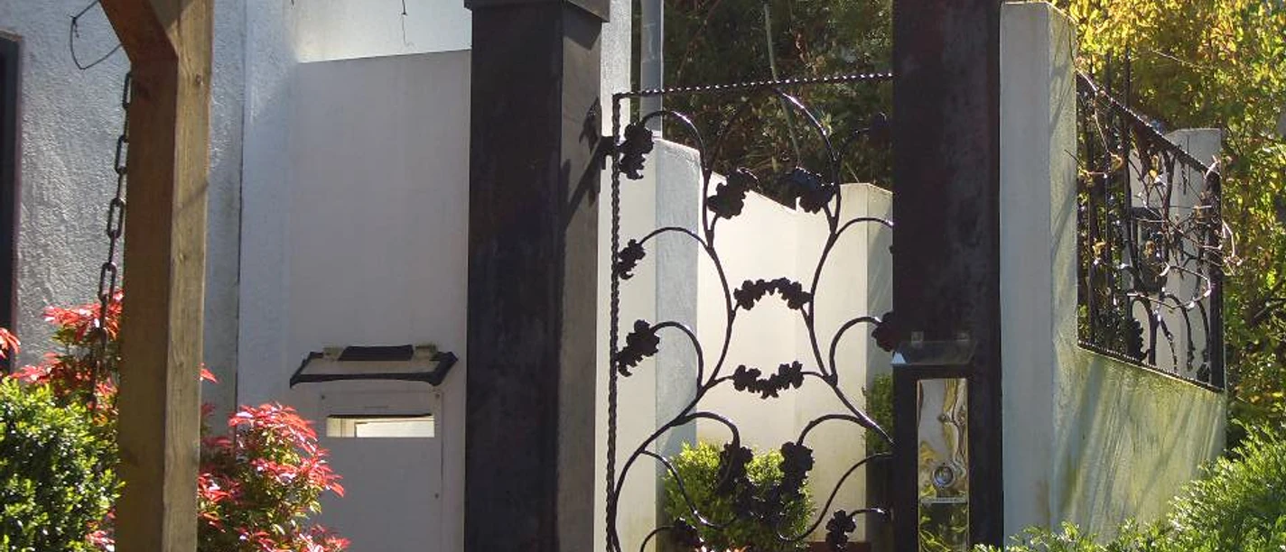 Gartenansicht Zierliches Gartentor aus Metall mit kunstvollem Rankendekor, flankiert von Pflanzen in Sonne.