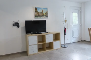 Ferienwohnung Berkenroth Ein heller Raum mit grauem Fliesenboden, Fernseher auf Regal, Gemälde an der Wand und Tür rechts.