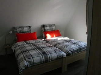 Zwei Einzelbetten mit karierter Bettwäsche und roten Kissen in einem hellen, minimalistischen Raum.