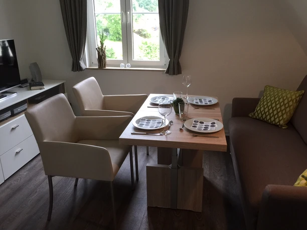 Ferienwohnung Buchenweg Gemütliches Wohnzimmer mit Esstisch, gedeckt für vier Personen, und zwei bequemen Sesseln in moderner Einrichtung.