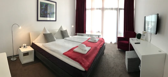 Gästehaus beim Prinz Helles Hotelzimmer mit Doppelbett, rot-weißer Bettwäsche, Schreibtisch, Fernseher und großem Fenster.