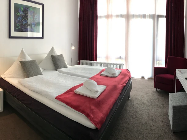 Gästehaus beim Prinz Helles Hotelzimmer mit Doppelbett, rot-weißer Bettwäsche, Schreibtisch, Fernseher und großem Fenster.
