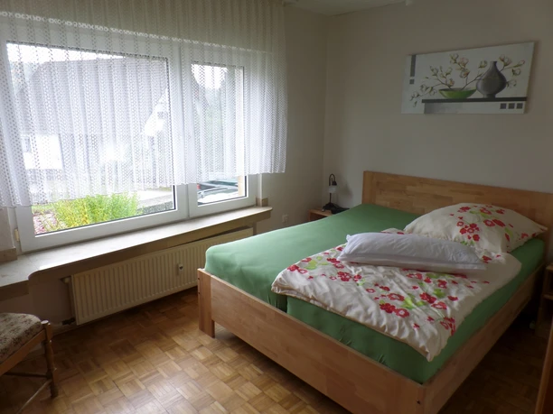 Ferienwohnung in Miebach Helles Schlafzimmer mit Holzparkett, einem Bett mit grünem Bettbezug und floralem Kissen, daneben ein Fenster.