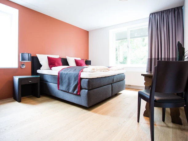 Hotelzimmer Modernes Hotelzimmer mit Doppelbett, roten Akzenten, Holzfußboden und zwei großen Fenstern.