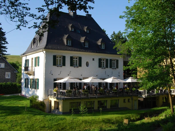 Außenansicht Historisches zweistöckiges Gebäude mit Schieferdach, Terrasse und Gästen, umgeben von Bäumen und Natur.