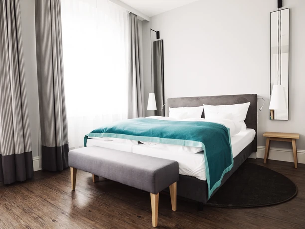 Doppelzimmer Modernes Hotelzimmer mit Doppelbett, türkisfarbenem Überwurf, Holzboden, hellen Vorhängen und Beleuchtung.
