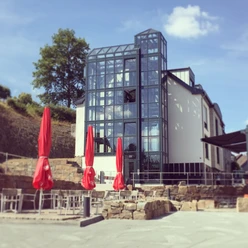 Außenansicht Modernes Gebäude mit großer Glasfassade, umgeben von Terrassenbestuhlung und roten Sonnenschirmen.