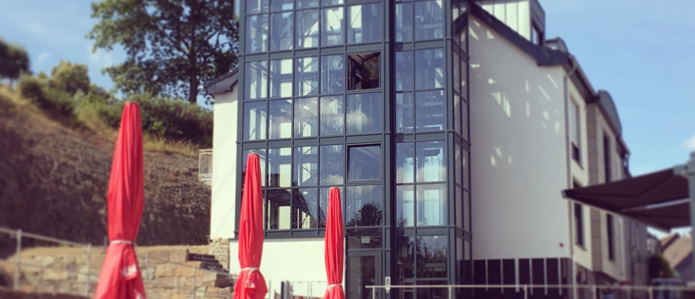 Modernes Gebäude mit großer Glasfassade, umgeben von Terrassenbestuhlung und roten Sonnenschirmen.