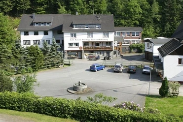 Haus am Mühlenberg Gasthof in ländlicher Umgebung mit Parkplatz, umgeben von Bäumen, und einem Brunnen im Vordergrund.