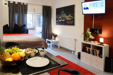 Wohnraum Wohnung A Modernes Apartment mit Doppelbett, TV, Esstisch, Obstschale und Stadtbild an der Wand.