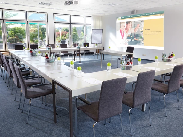 Tagungsraum Heller Seminarraum mit U-förmiger Tischanordnung, Sitzplätzen, Notizmaterial und begrünten Fenstern.