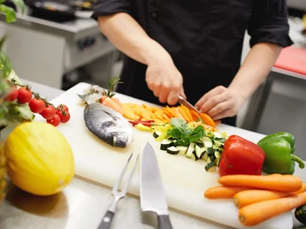 Küche Koch schneidet Gemüse neben frischem Fisch in einer hellen, modernen Küche auf einem Schneidebrett.