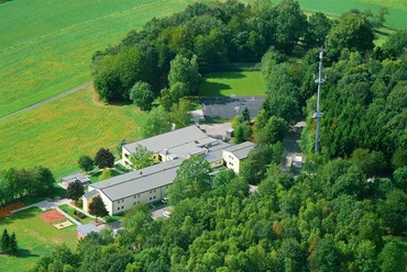Hotel Fit Luftbild Luftaufnahme eines Gebäudekomplexes inmitten grüner Wälder und Felder, mit Turm im Hintergrund.