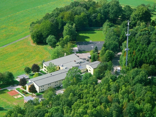 Hotel Fit Luftbild Luftaufnahme eines Gebäudekomplexes inmitten grüner Wälder und Felder, mit Turm im Hintergrund.