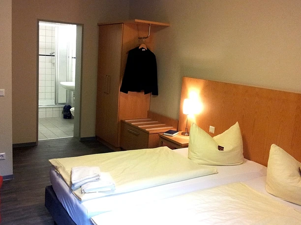 Hotel Fit Zimmerbeispiel Ein schlichtes Hotelzimmer mit Doppelbett, offenen Kleiderschrank und angrenzendem Badezimmer.
