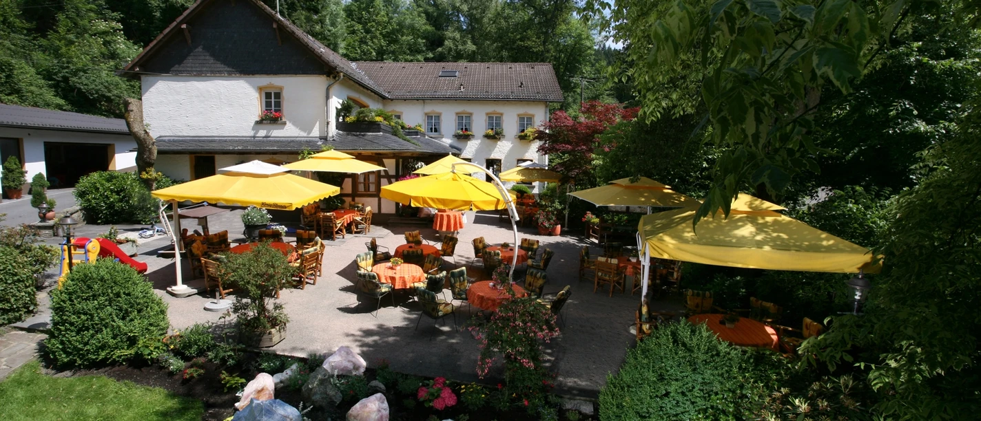 Hotel Rengser Mühle Ein idyllischer Gastgarten mit gelben Schirmen und Tischen vor einem charmanten zweistöckigen Gebäude.