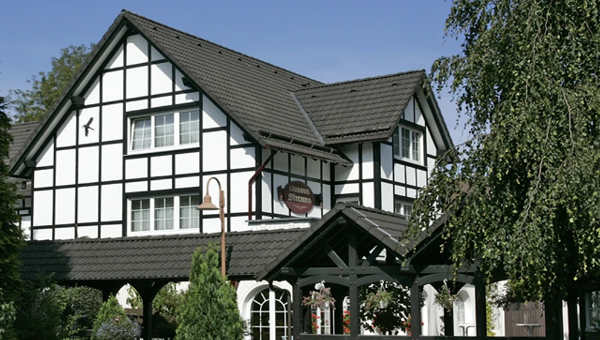 Außenansicht Fachwerkhaus mit schwarzem Holzrahmen, dekorativen Fenstern und grünem Garten unter blauem Himmel.
