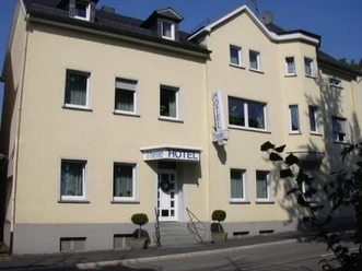 Gelbes, dreistöckiges Gebäude mit weißem Schriftzug „Hotel" über dem Eingang, umgeben von Bäumen.