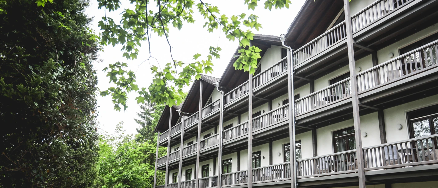 Außenansicht Mehrstöckiges Gebäude mit Holzbalkonen, umgeben von grünen Laubbäumen, unter blauem Himmel.