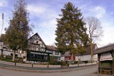 Außenansicht Fachwerkhaus mit Restaurant-Pension, umrahmt von Bäumen, unter blauem Himmel an einer Landstraße.