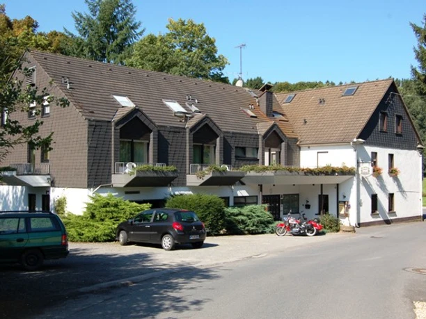 Hausansicht Ein zweigeschossiges Gebäude mit dunkler Schieferfassade, geparkten Autos und umgeben von Bäumen.