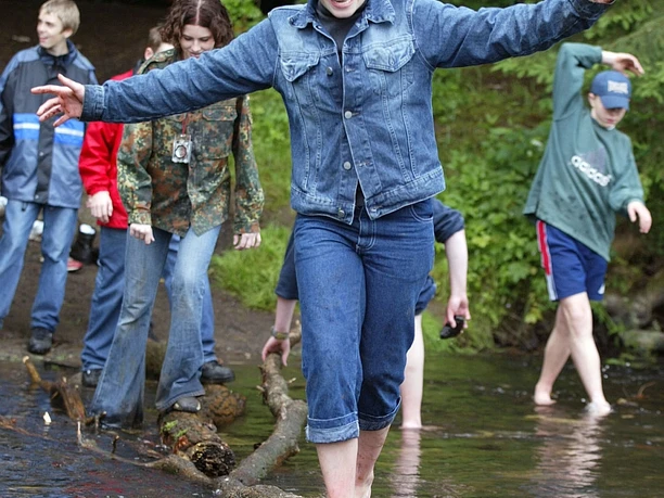 JHB Wipperfürth Kinder balancieren barfuß über einen Baumstamm in einem Bach, umgeben von grüner Natur.