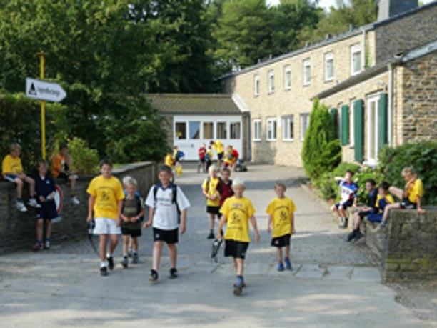 Jugendherberge Wipperfürth Kinder in gelben T-Shirts laufen und stehen auf einem gepflasterten Hof vor einem großen Gebäude.