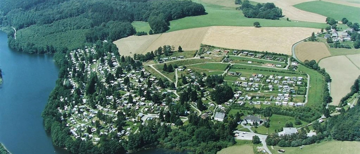 Campingplatz Lambacher Höhe Luftaufnahme eines Campingplatzes und Grünflächen an einem Seeufer, umgeben von Wäldern und Feldern.