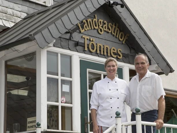 Landgasthof Tönnes Ein herzliches Paar vor dem Eingang eines charmanten Landgasthofs mit Holzverzierungen.