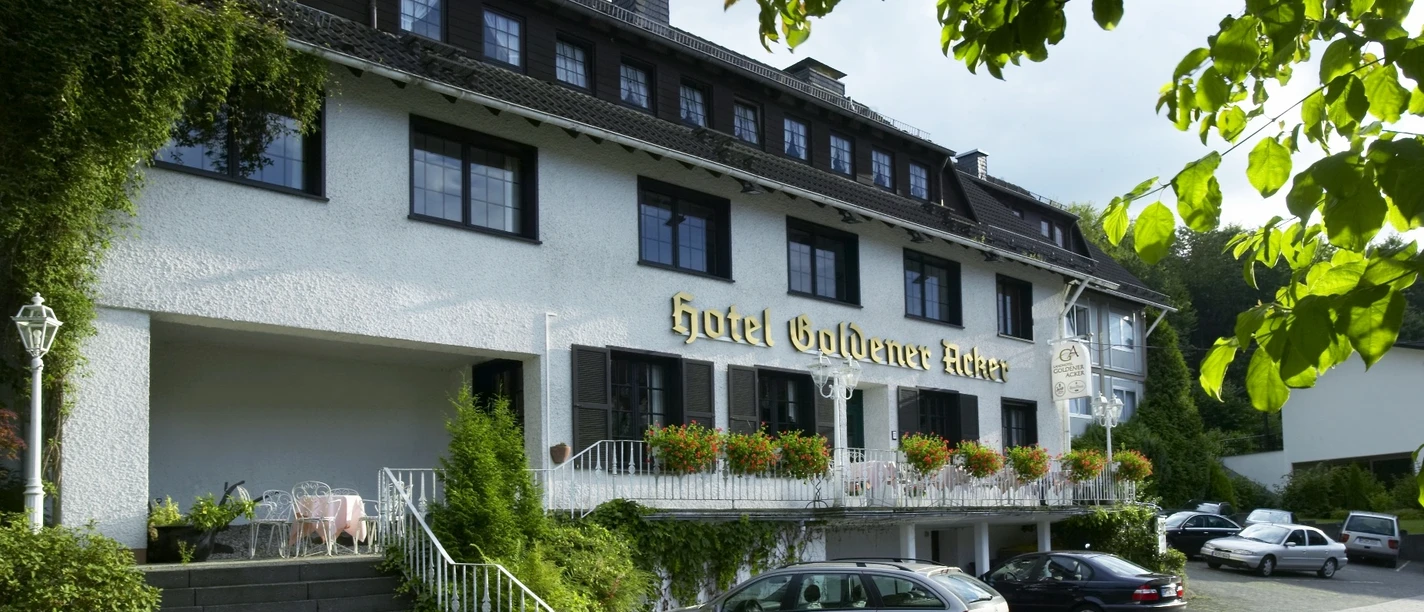 Landhotel zum goldenen Acker Hotelgebäude mit weißer Fassade, großem Balkon und blühenden Pflanzen, umgeben von geparkten Autos.