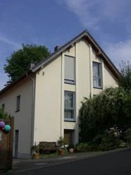 Lustig - Ferienwohnung Bensberg Einfamilienhaus mit Satteldach, gelber Fassade, großen Fenstern, umgeben von Pflanzen und Bäumen.