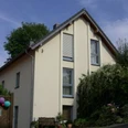 Lustig - Ferienwohnung Bensberg Einfamilienhaus mit Satteldach, gelber Fassade, großen Fenstern, umgeben von Pflanzen und Bäumen.