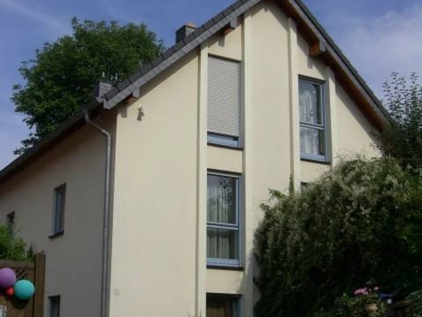 Lustig - Ferienwohnung Bensberg Einfamilienhaus mit Satteldach, gelber Fassade, großen Fenstern, umgeben von Pflanzen und Bäumen.