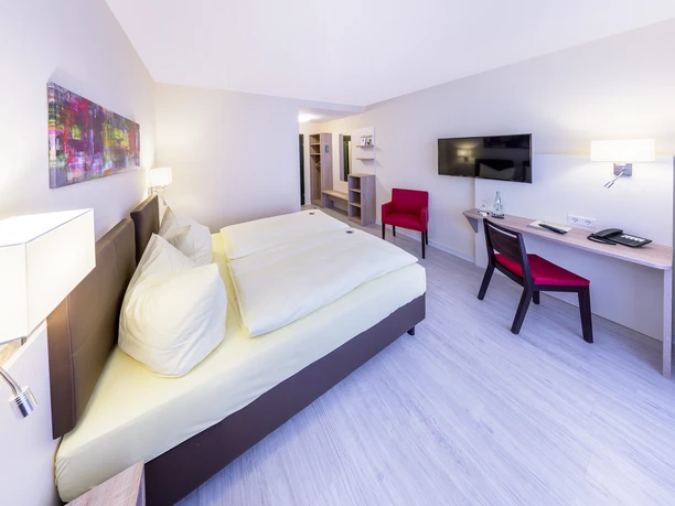 Montana Hotel Gummersbach Nord Modernes Hotelzimmer mit Doppelbett, Schreibtisch, rotem Stuhl, Wandkunst und Flachbildfernseher.