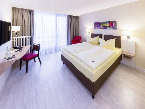 Montana Hotel Gummersbach Nord Ein modernes, helles Hotelzimmer mit Doppelbett, Schreibtisch, Flachbildfernseher und Sessel.