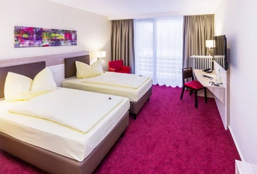 Montana Hotel Gummersbach Nord Hotelzimmer mit zwei Einzelbetten, rotem Teppich, TV, Schreibtisch und heller Fensterfront.